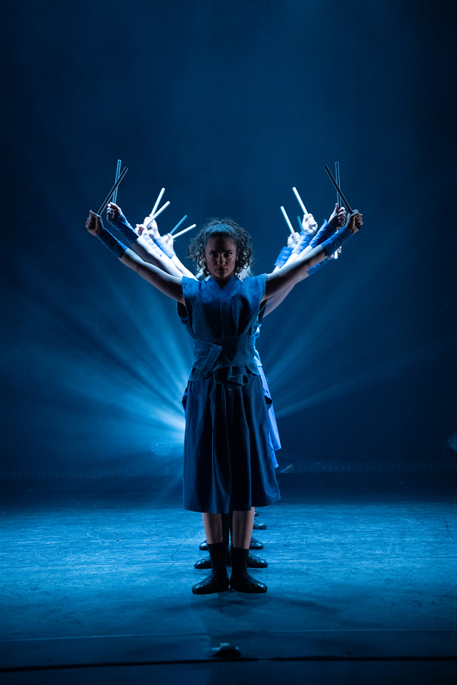 Six woman dancers bathed in blue light in a downstage to upstage line hold sticks aloft on either sides of their bodies
