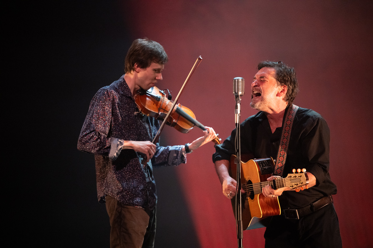 A guitartist and a fiddler face one another. Both are playing and the guitarist is singing into a standing mic.