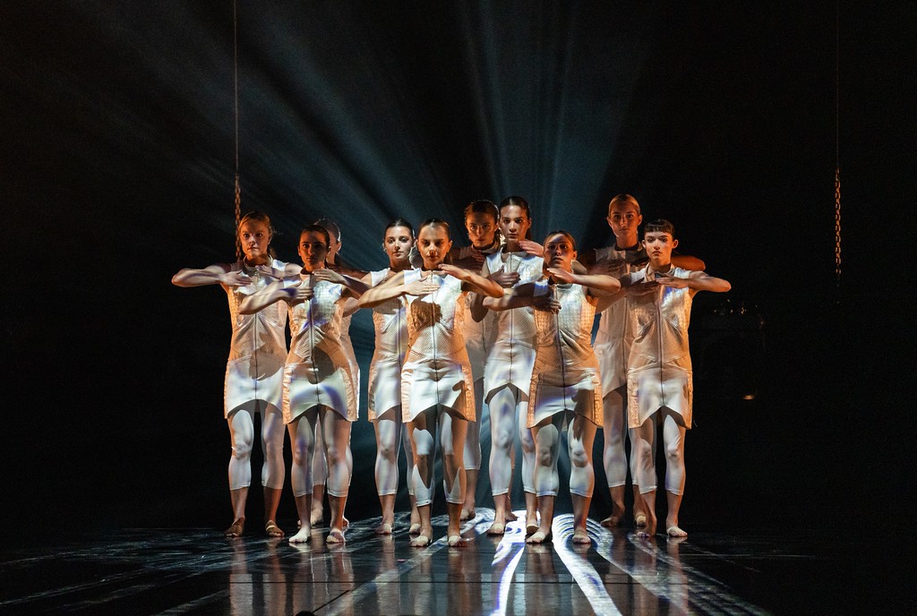 A tightly spaced group of women dressed in sleek white dresses and leggings, feet bare, face the audience with arms bent at the elbow chest high.