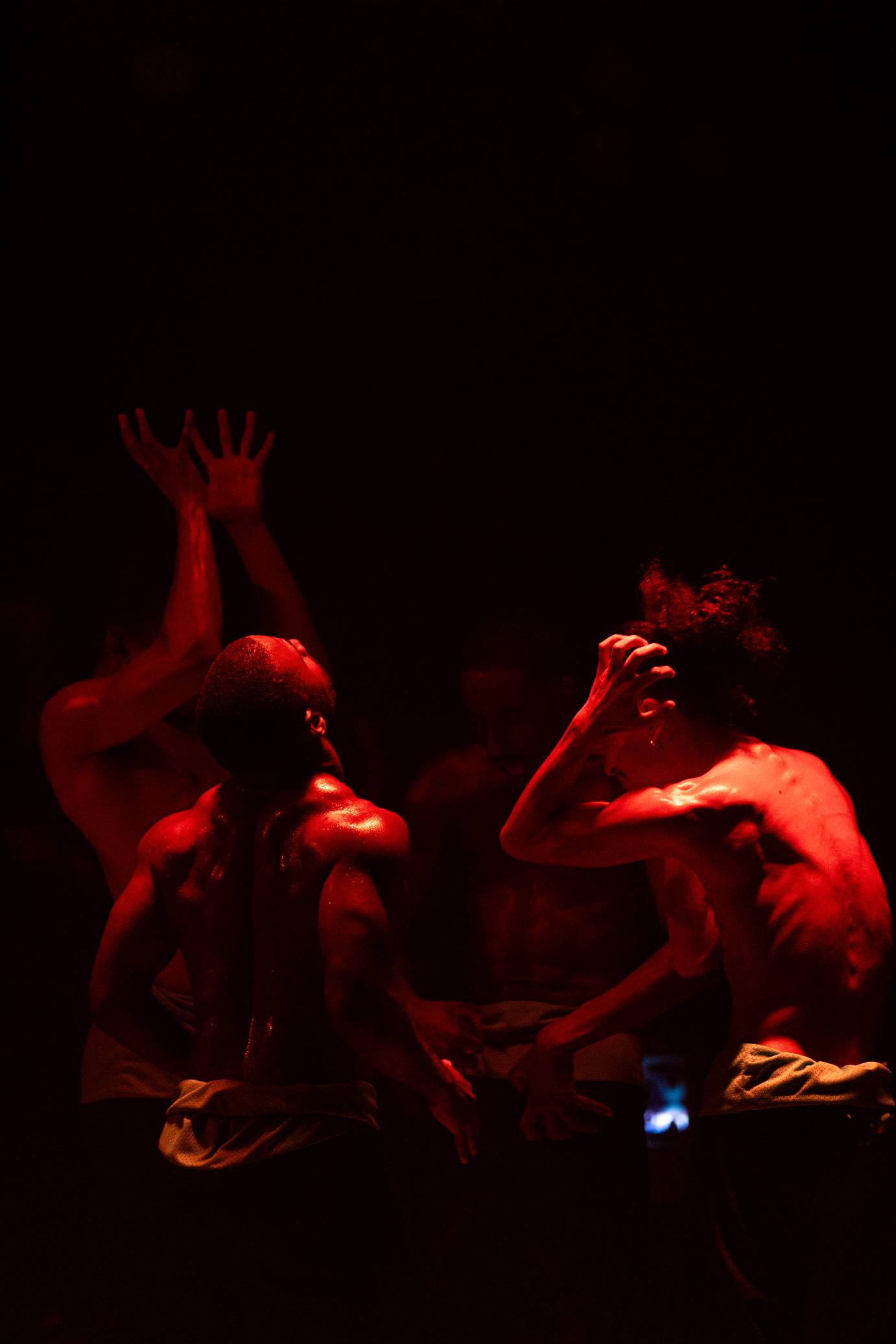 Three male, sweaty and muscular torsos, bathed in red light, express tension in clenched fingers, an upturned face and arms extended overhead with fingers splayed.