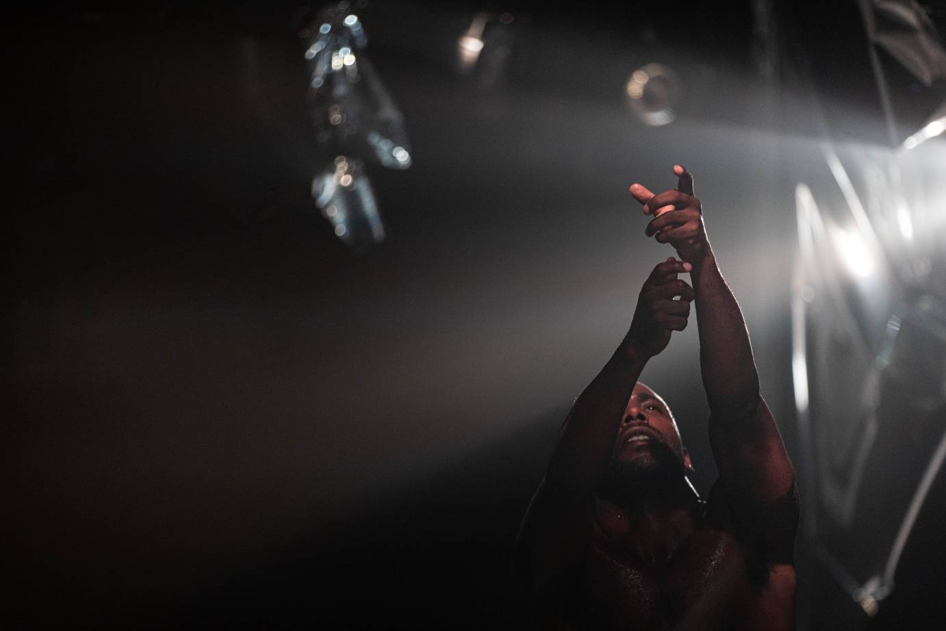 A man's face, partially obscured by two uplifted arms with pointed fingers, is illuminated by a white side light.