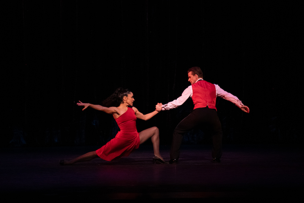 a woman in a bold red off the shoulder dress stares at her partner in a red vest and tuxedo pants. They are in oppostion, she on one knee pulling away from him but looking interested in getting together and he holding her hand looking as if he is ready to pull her to him at any minute 