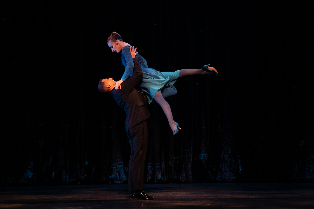 a man in a tuxedo lifts his female partner high in the air as she faces him and holds on to him. She is dressed in an aqua silk short dress. 