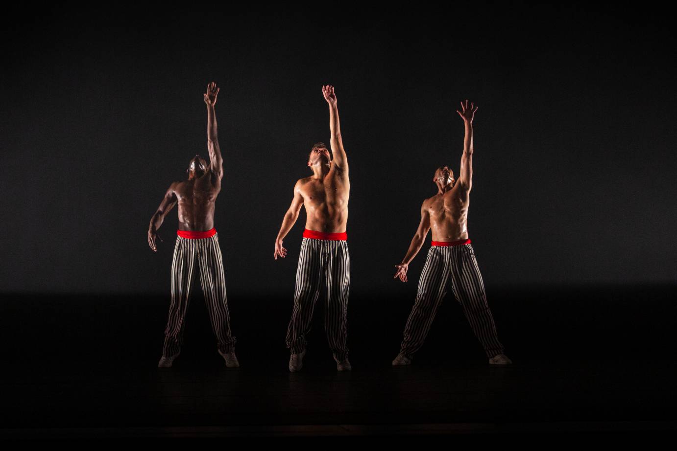 three bare chested men, on the tips of there toes look upwards, streching one arm to the sky. The wear silky black and white pants and white sneakers, and are covered in sweat