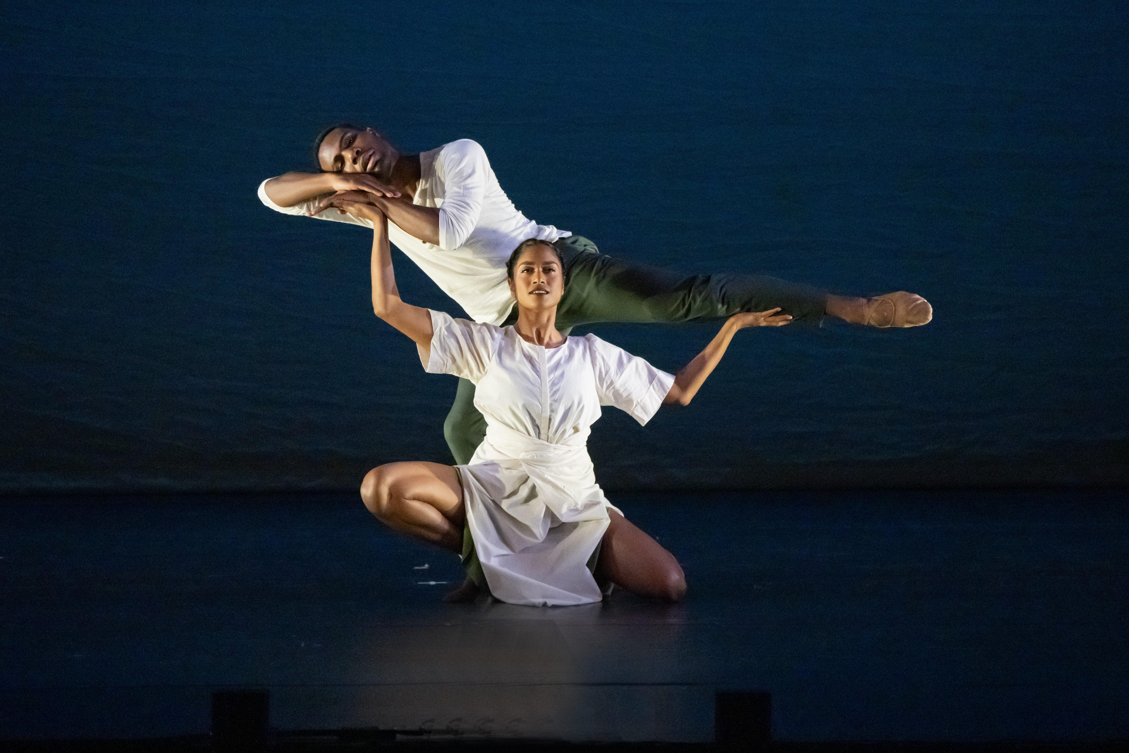 female dance kneels, hands up, supporting the arms and leg of a  Male dancer 