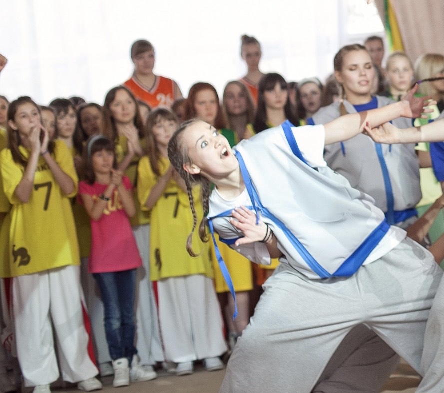 a girl with wide eyes, braids, performing admist a crowd of clapping children