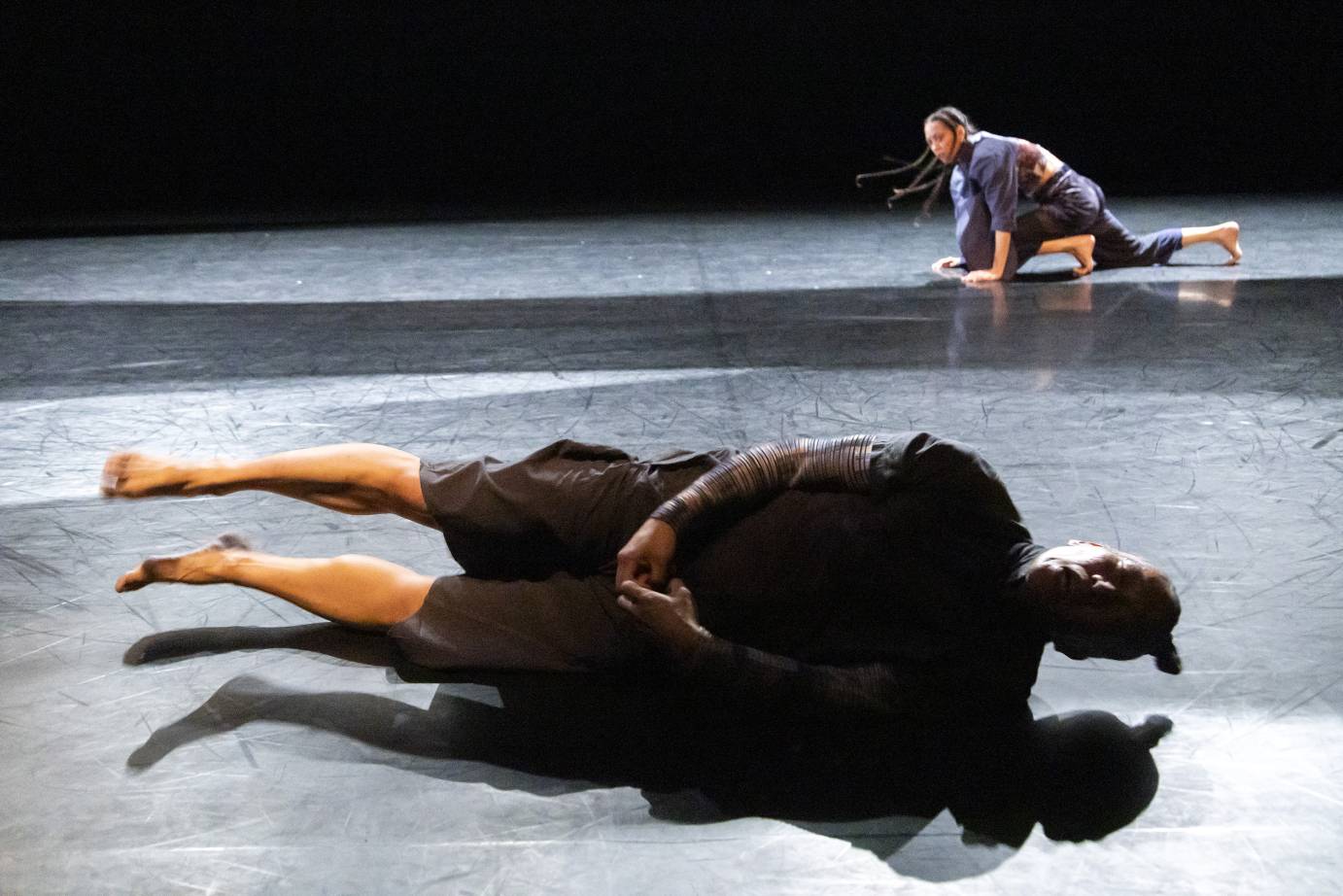 A man clothed in gray perches on the side of his body as if he were rolling downstage; hands are clasped in front. A woman with long braids lunges far upstage.