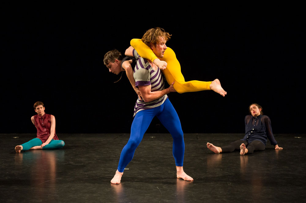 2 dancers sit casually on a marley looking at a central couple consiting of a man in a white and purple striped t shirt with tight blue leggings holding a person in bright yellow tights on his shoulders. the yellow-tighted person looks as if they is climbing up the man carrying them.