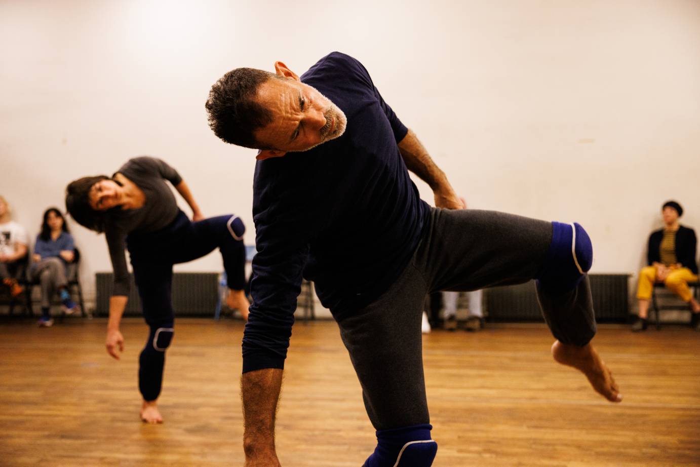 two dancers on a wooden floor both clad in black and both  standing on one leg in a side tilt, with their free leg bent 
