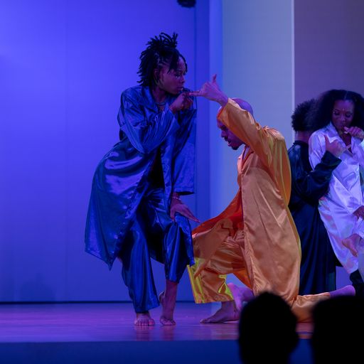 Four dancers near the stage entrance: the dancer most downstage, costumed in salmon, on bended knee with gaze lowered and left arm raised above head. The dancer to their right, costumed in midnight blue, points to the two exiting dancers