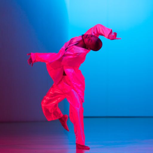 Man with head and right arm thrown up and back, dressed in flowing satin hot pink tunic and pants against a bright blue background