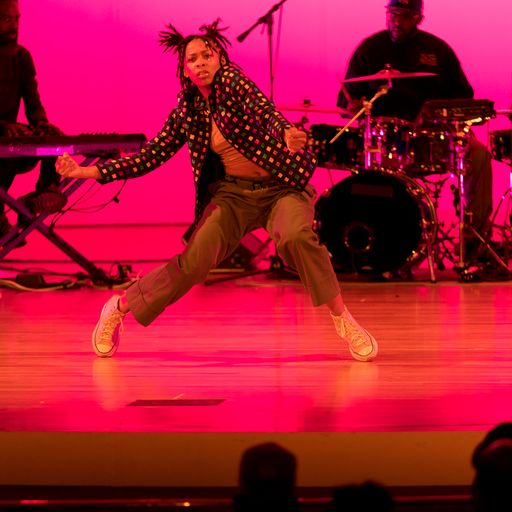 Woman dancer with two pigtails on top of her head, shrugs her shoulders, on tip toes with knees bent. Dressed in a loose polka dot shirt and dark pants