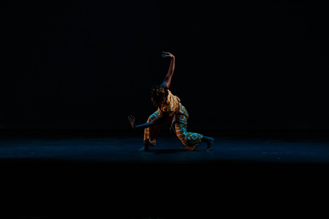 A dancer, dressed in a colorful orange and white costume, deeply lunges with one arm stretched upward.