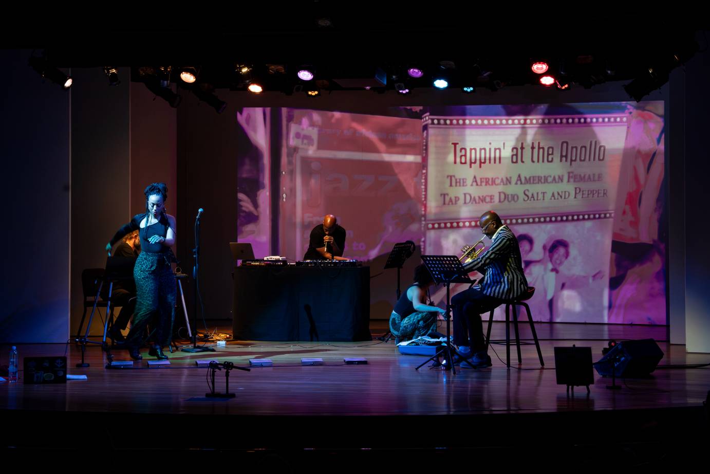 The upstage screen, bathed in purple, acts as the backdrop for a standing woman how looks down and a seated trumpeter.