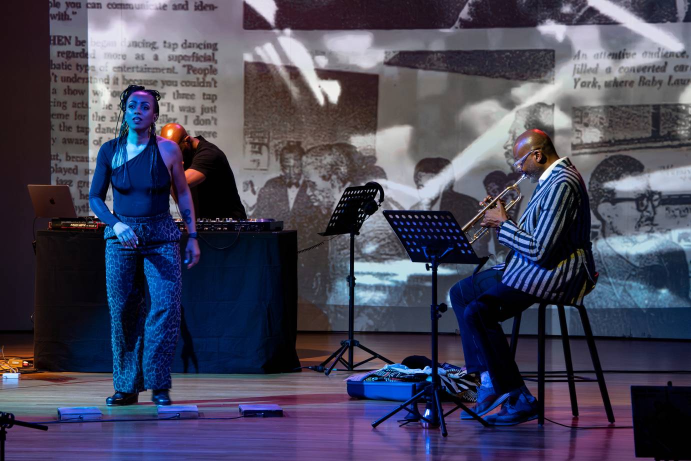 A woman tapper looks out to the audience while the trumpet player, dressed in a striped jacket, looks down at his sheet music. Images of faces are screened behind them.