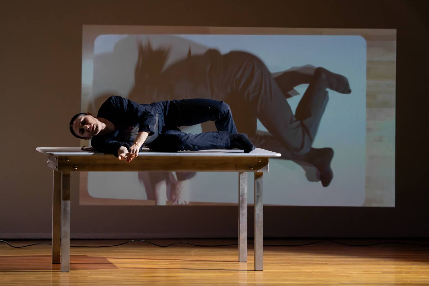 A woman lies on her side facing out on a small table. Her image is projected behind her on a white screen.
