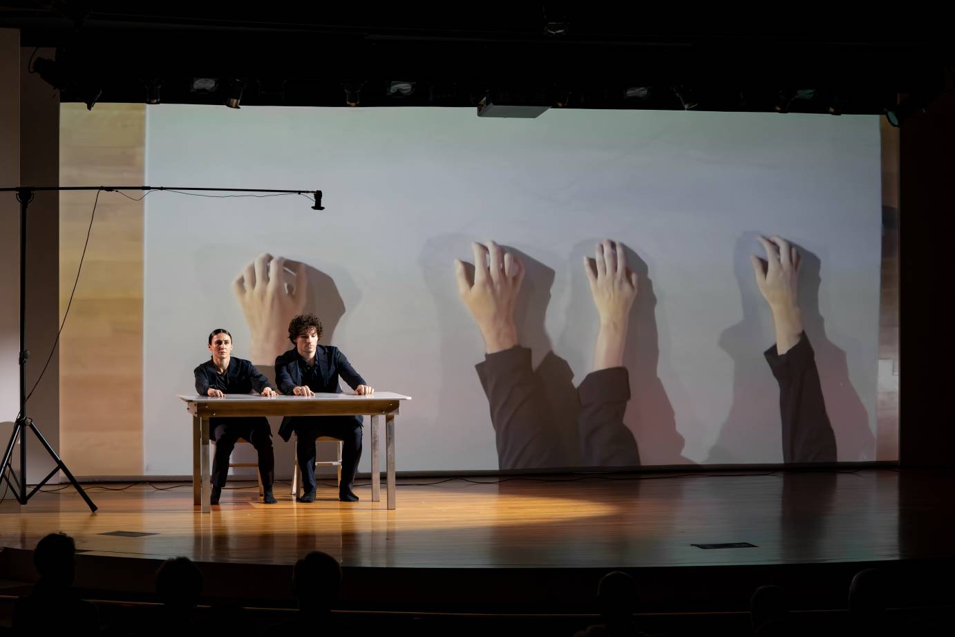 A woman and a man dressed in dark clothing sit close to one another their hands resting on the top of the table. Concurrently, their hands, with fingers pointed upward, are projected behind them.