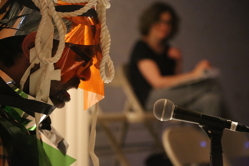 Brian Bukhari is shrouded in rope, paper and material and sits in front of a microphone