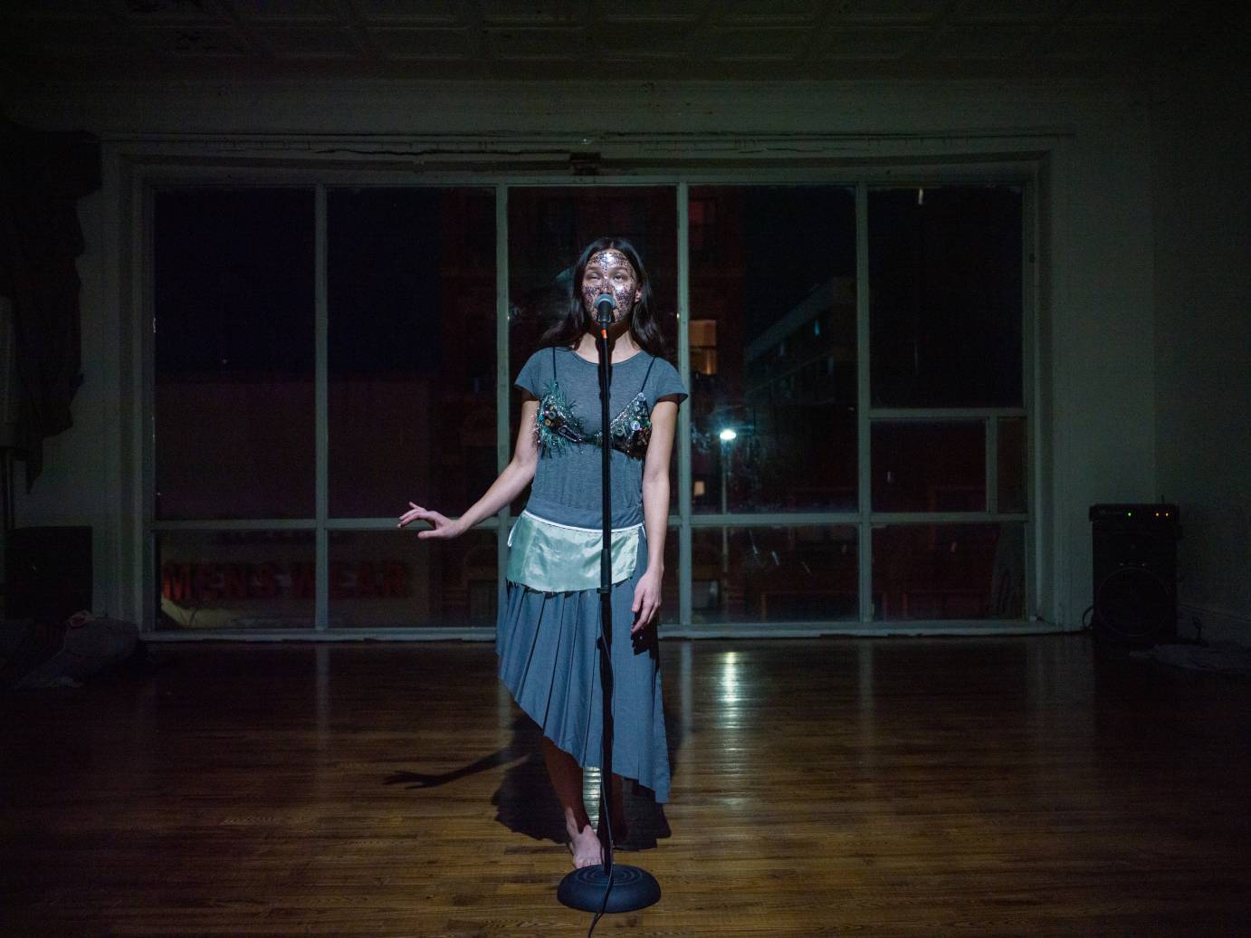 a singer at a standing microphone wearing  a grey dress with glitter bra and tiny glitter apron 