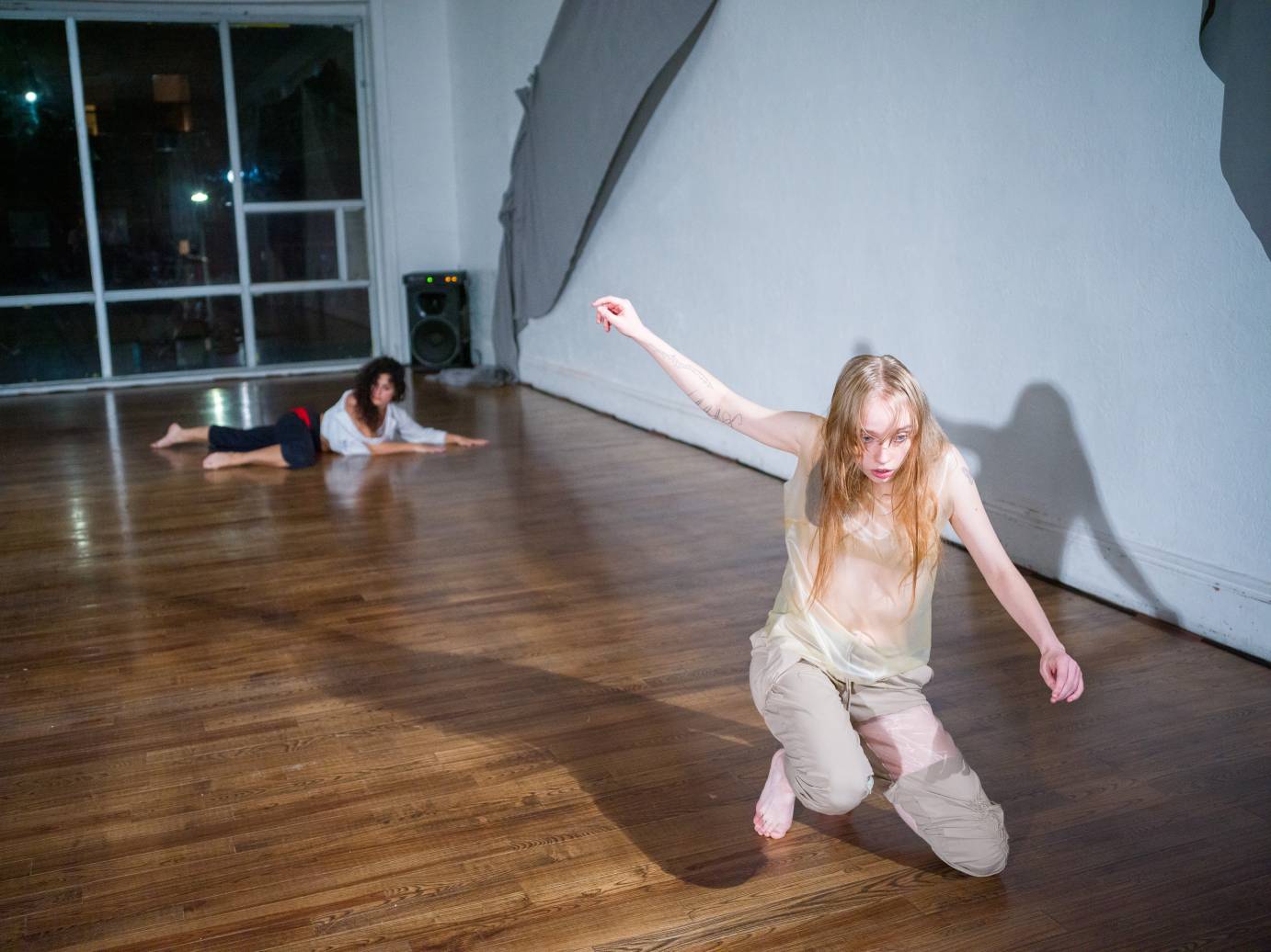 one woman in mesh kneels on the floor, looking surprised or  poised to attack.. behind her another dancer in a sweat outfit of dark pants and grey shirt, seems to be crawling along the floor 