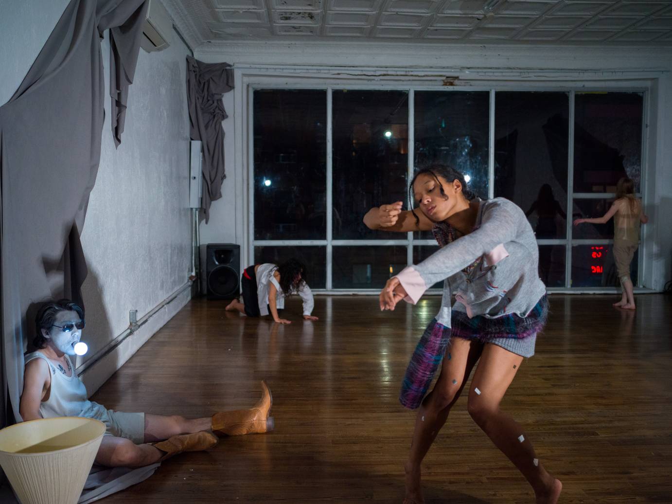 young dancers arranged around a room with large windows at its back, wearing  a combination of sweats, mesh, and rags, all seem in their own world. a person whose face is painted blue has a lit lightbulb in his mouth 