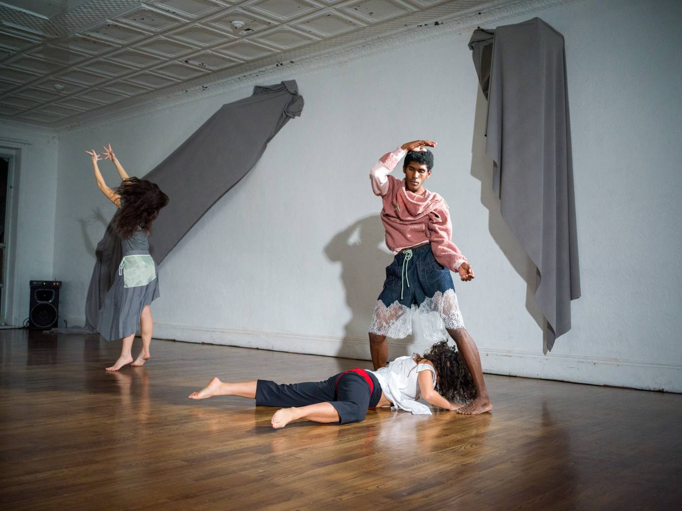 a trio of dancers wearing loose sweats of different colors, they move in front of a white wall from which two grey swaths of cloth hang