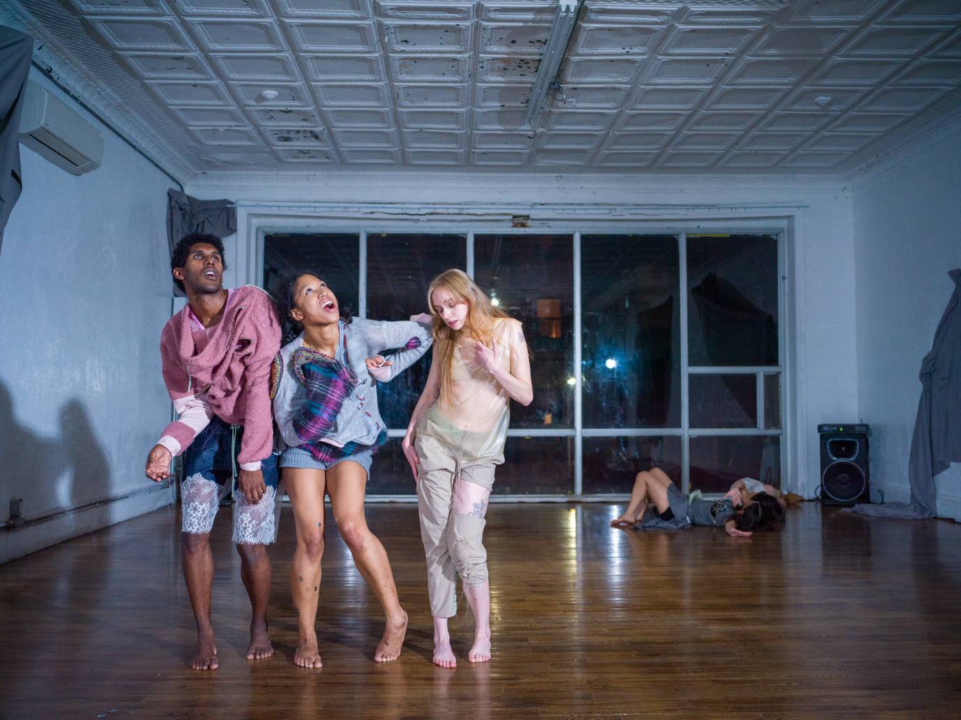 three dancers stand pressed together and hunched in posture while in the background of the windowed studio two dancers like on the floor