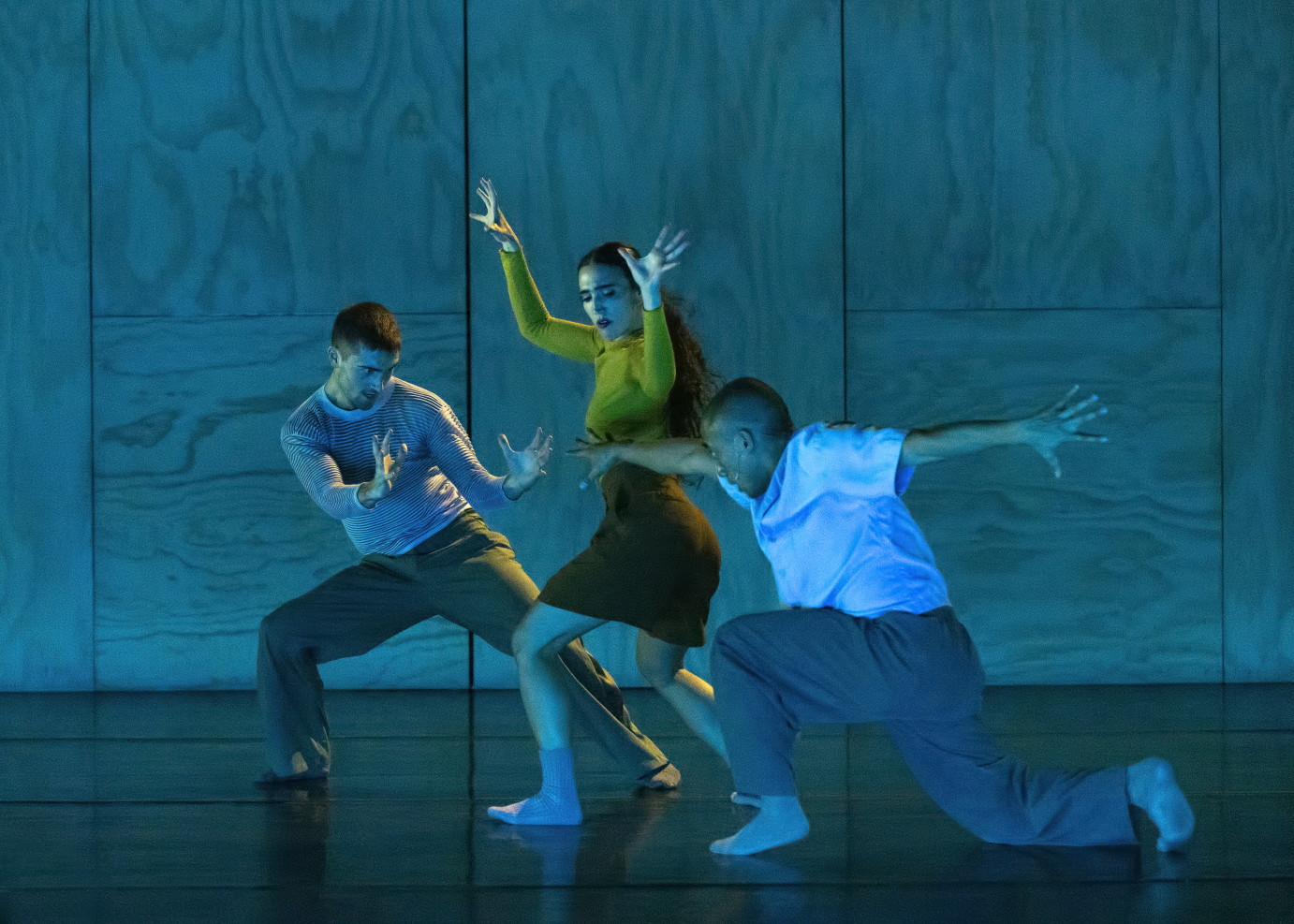 Two men lunge around a woman in a yellow top. She has her arms lifted and bent at right angles at the elbow.