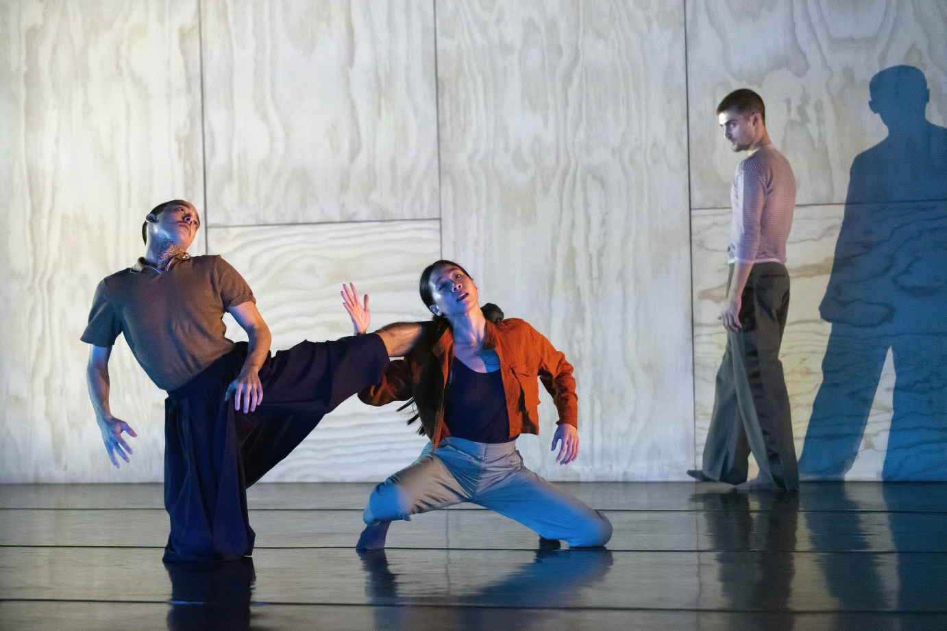 Against a plywood wall, a kneeling woman holds up the leg of a man. Another man looks on in profile.